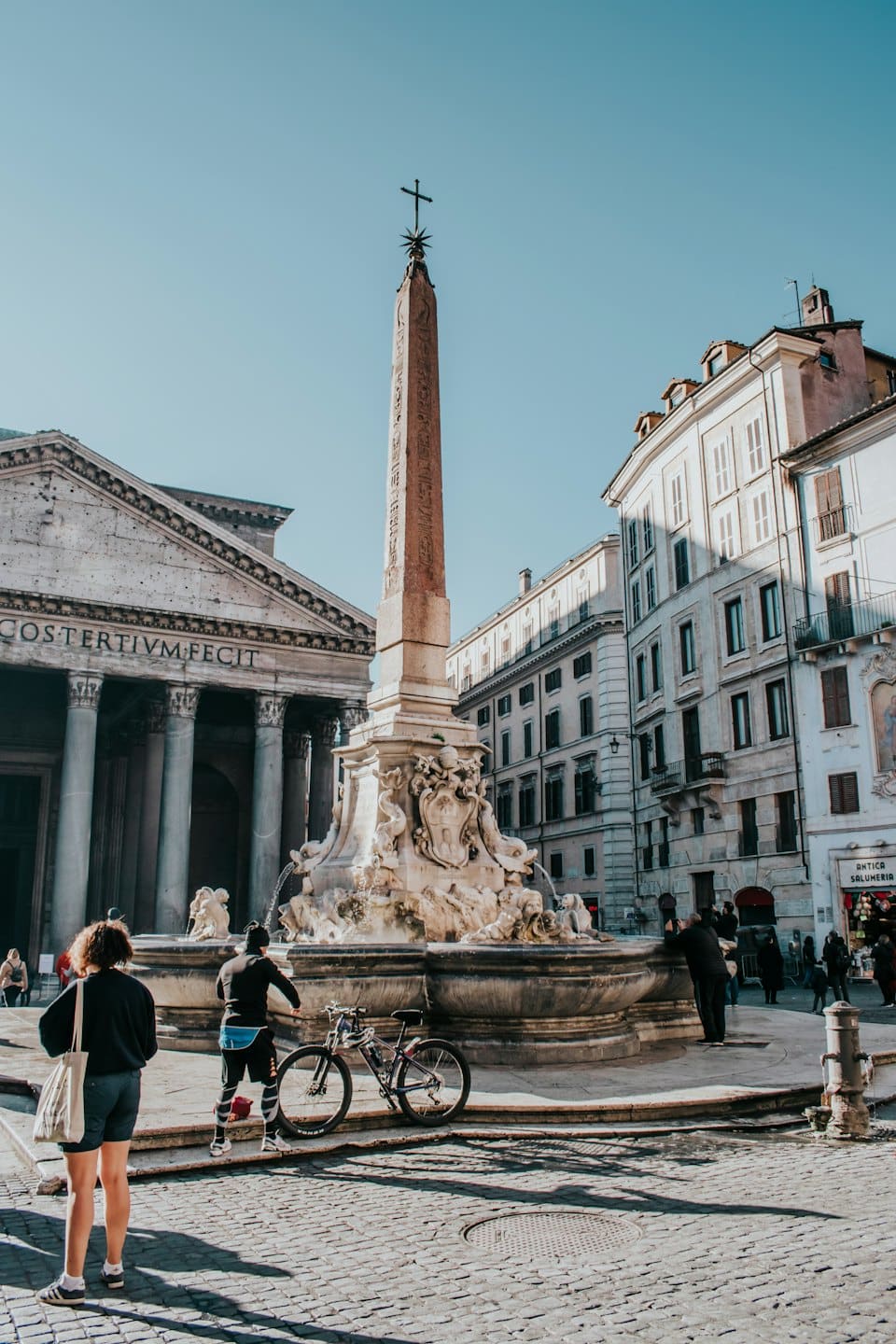 Persone riunite attorno alla fontana del Pantheon in una piazza romana soleggiata.