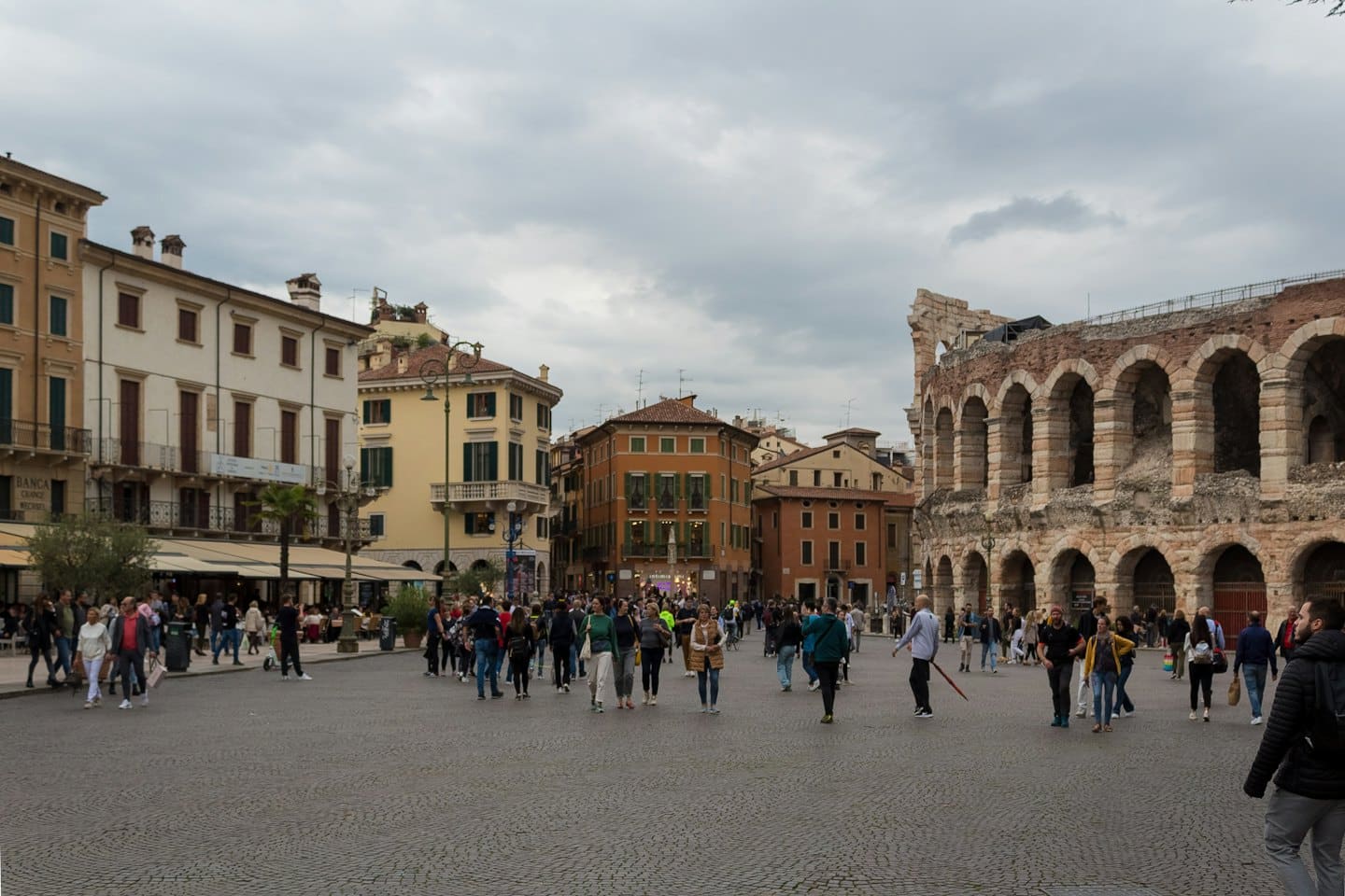 Persone passeggiano in una piazza europea affollata con anfiteatro e palazzi antichi.